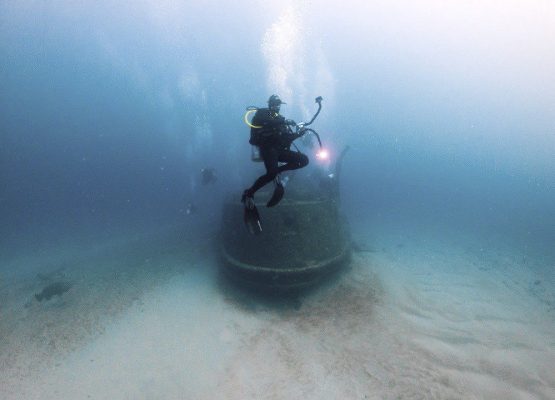 Alain scuba diving in the ocean with a wreck behind him.