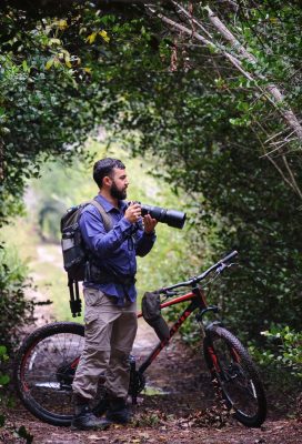 Alain standing in a forest with a mountain bike behind him and a large camera in his hand.